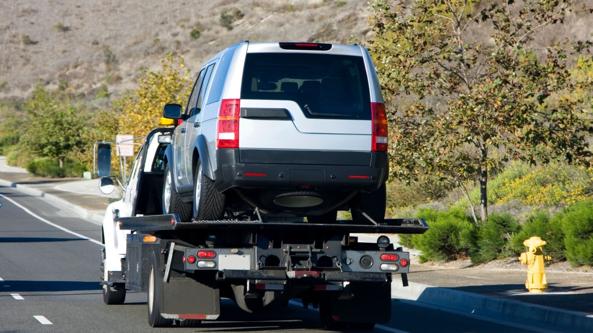White SUV being towed on a flatbed truck along a road with trees